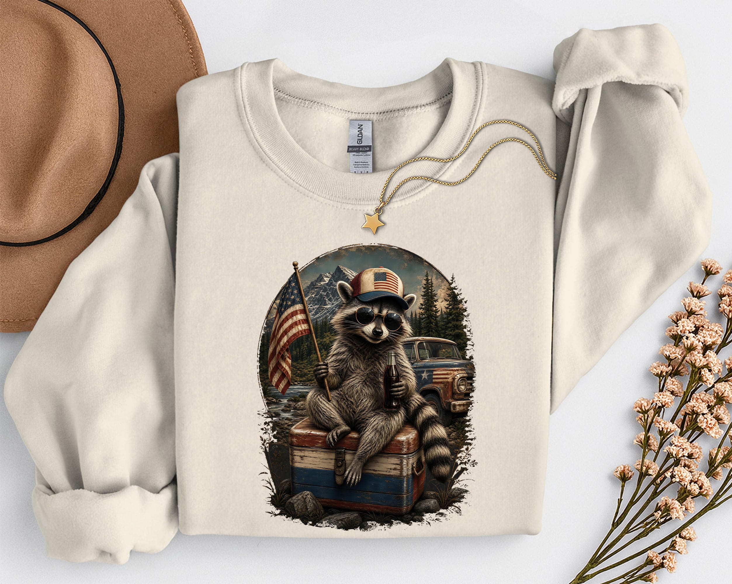 raton-laveur-usa-sand Un sweat-shirt sable à manches longues orné d'un motif représentant un raton laveur portant des lunettes de soleil et une casquette aux couleurs du drapeau. Il est assis sur une glacière, tenant un petit drapeau américain et une bouteille.