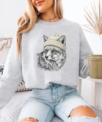Femme portant un sweat gris clair avec un renard en bonnet illustré au crayon, tasse de café à la main