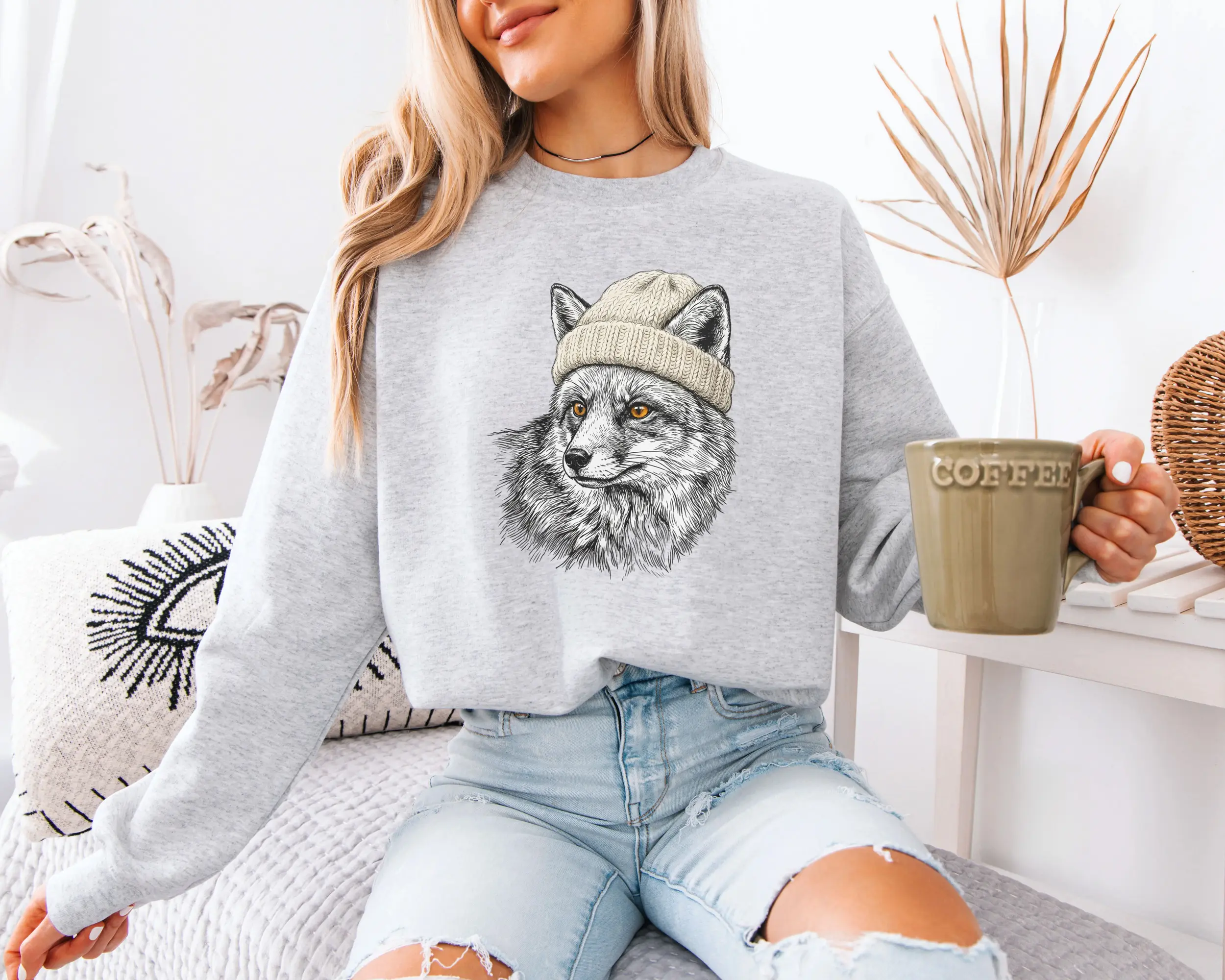 sweat-renard-bonnet-ash-f Femme portant un sweat gris clair avec un renard en bonnet illustré au crayon, tasse de café à la main